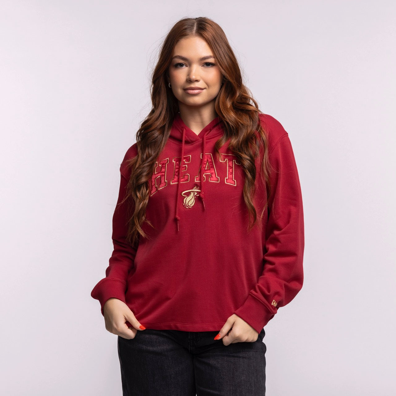 A woman with long brown hair wears the New Era Miami HEAT Open Back Women's Hoodie, featuring a basketball logo. This relaxed-fit, cropped cotton hoodie by New Era has a laid-back vibe and front pocket. Machine wash cold recommended.