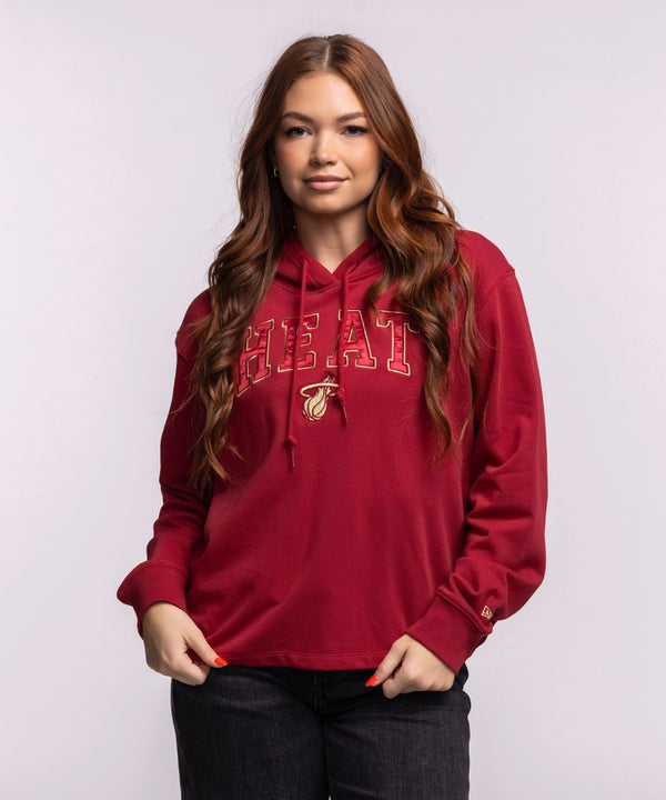 A woman with long brown hair wears the New Era Miami HEAT Open Back Women's Hoodie, featuring a basketball logo. This relaxed-fit, cropped cotton hoodie by New Era has a laid-back vibe and front pocket. Machine wash cold recommended.