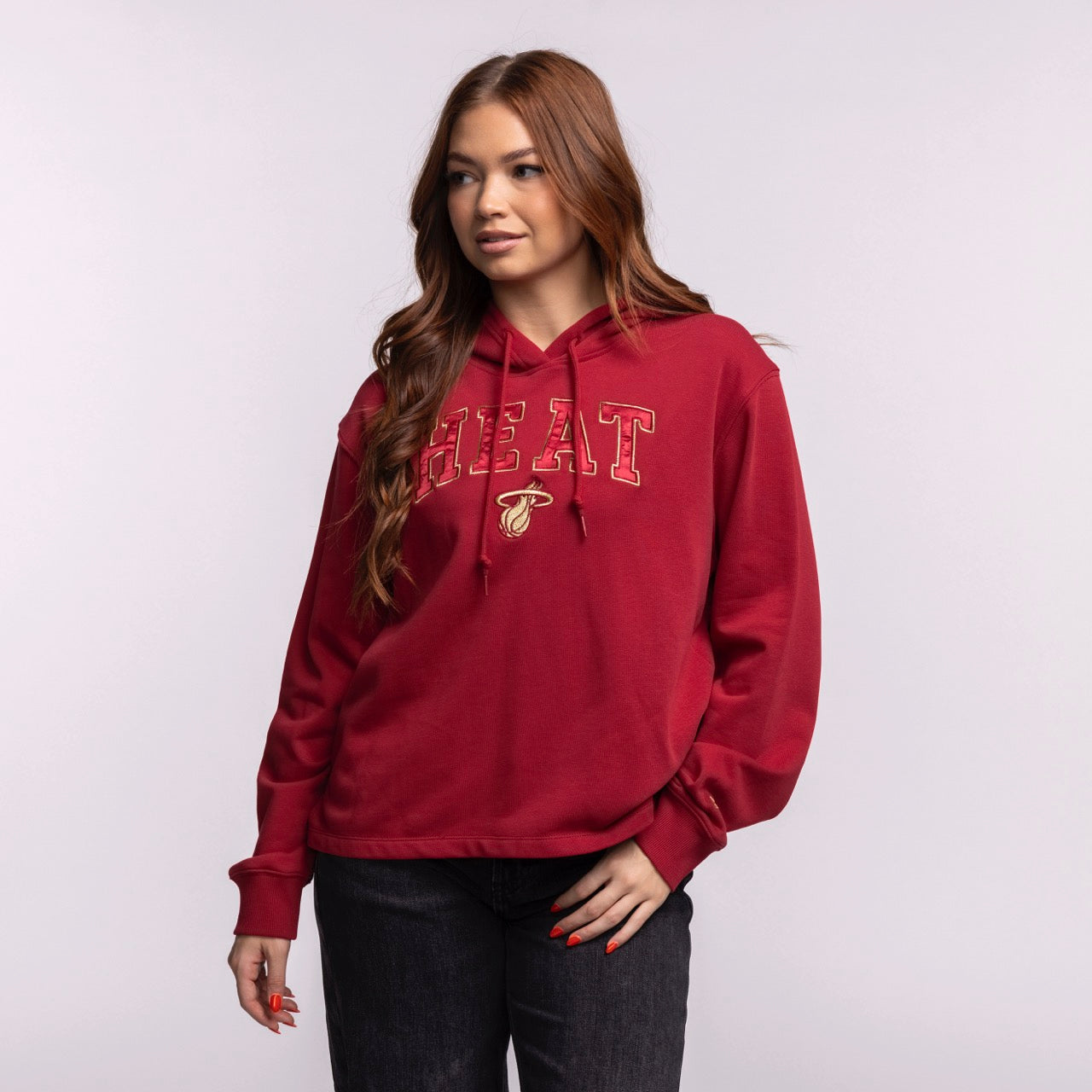 A woman with long brown hair models the New Era Miami HEAT Open Back Women's Hoodie in red and relaxed fit, paired with black jeans. She stands against a plain light gray background, gazing slightly to the side with one hand by her hip.