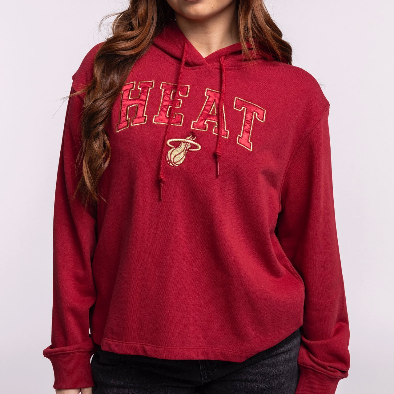 A woman wearing the New Era Miami HEAT Open Back Women's Hoodie, featuring a relaxed fit, red color, and team logo on the front, stands against a plain light background.