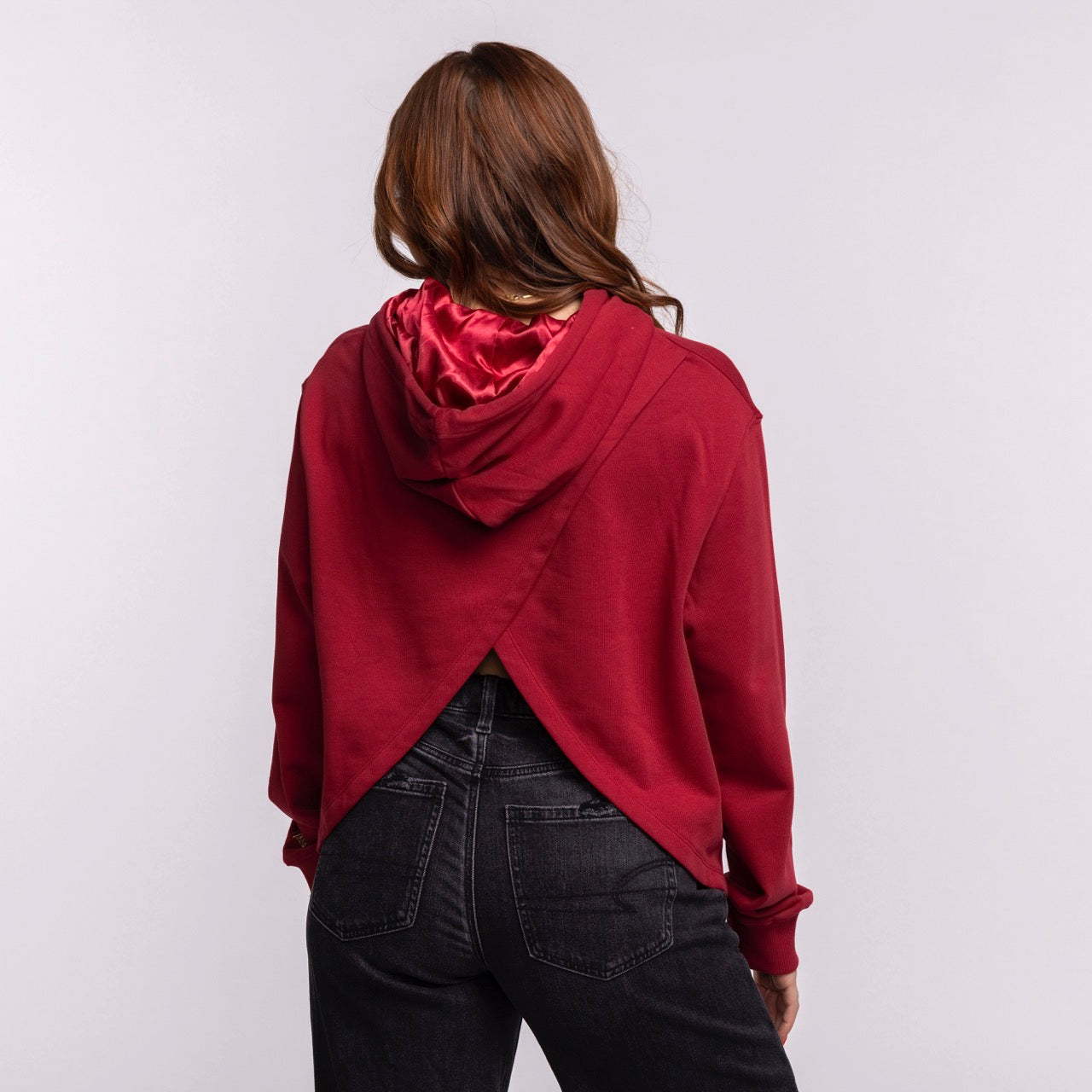 A woman with long brown hair wears the New Era Miami HEAT Open Back Women's Hoodie, featuring a relaxed cropped fit, satin-lined hood, crisscross open back, paired with high-waisted black jeans against a plain light background.
