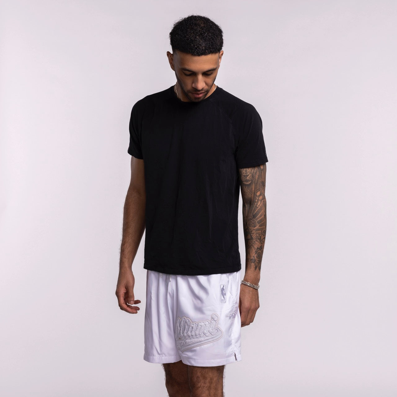 A man with short curly hair wears a plain black T-shirt and Pro Standard Miami HEAT White Out Woven Shorts. He stands against a light background, looking down with one arm relaxed and the other slightly bent, showing a tattoo on his forearm. Wash with like colors.