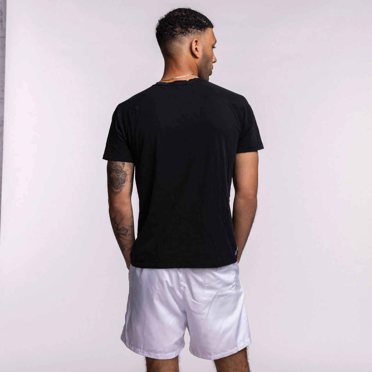 A man stands with his back to the camera, wearing a standard fit black t-shirt and Pro Standard Miami HEAT White Out Woven Shorts by Pro Standard. He has short hair, a tattoo on his left arm, and his hands relaxed against a plain light background.
