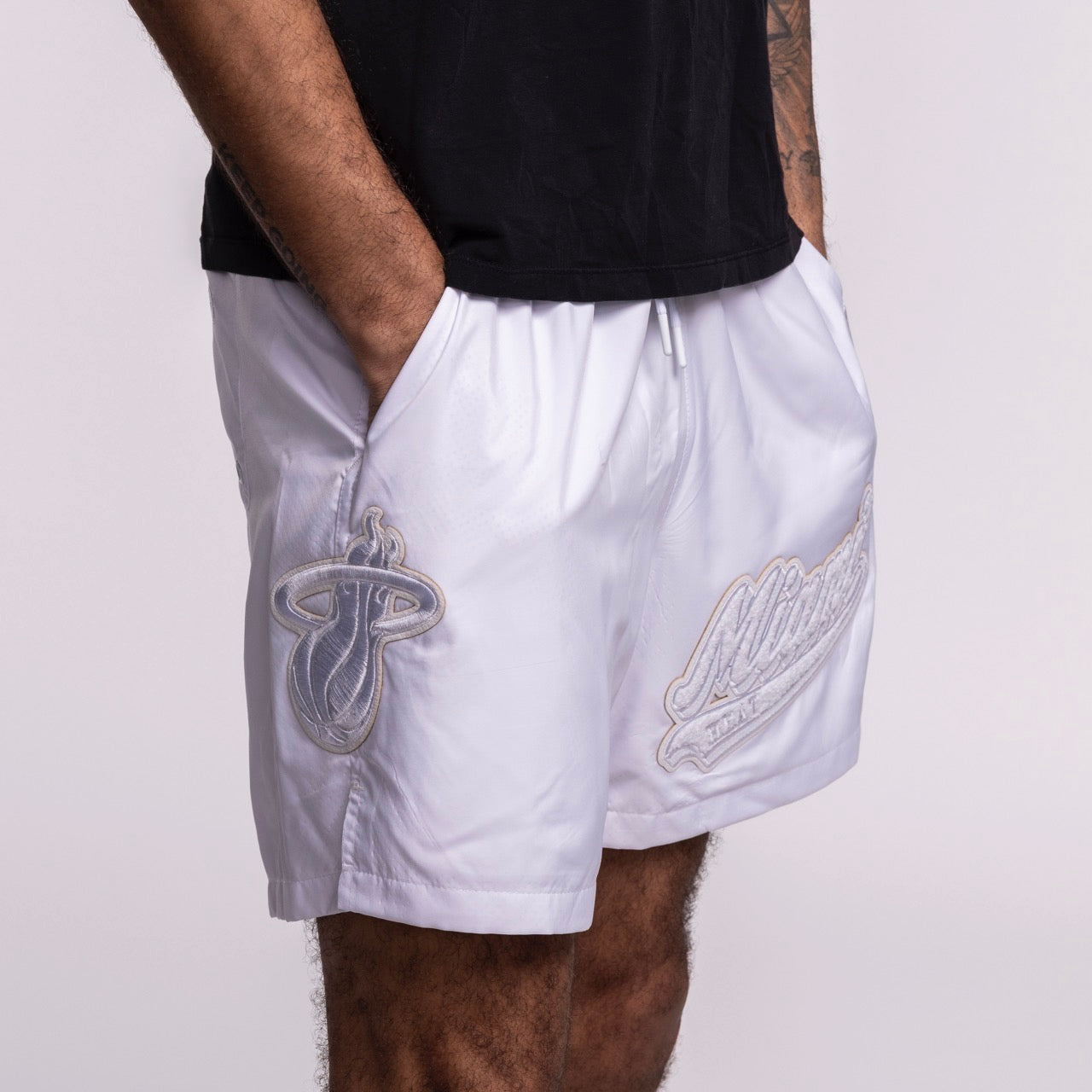 Wearing Pro Standard Miami HEAT White Out Woven Shorts, featuring Miami Heat and Milwaukee Bucks logos, hands in pockets, paired with a black t-shirt. Photo cropped mid-torso to mid-thigh. Wash with like colors for best care.