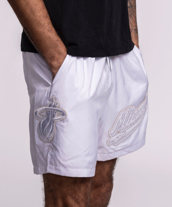 Wearing Pro Standard Miami HEAT White Out Woven Shorts, featuring Miami Heat and Milwaukee Bucks logos, hands in pockets, paired with a black t-shirt. Photo cropped mid-torso to mid-thigh. Wash with like colors for best care.