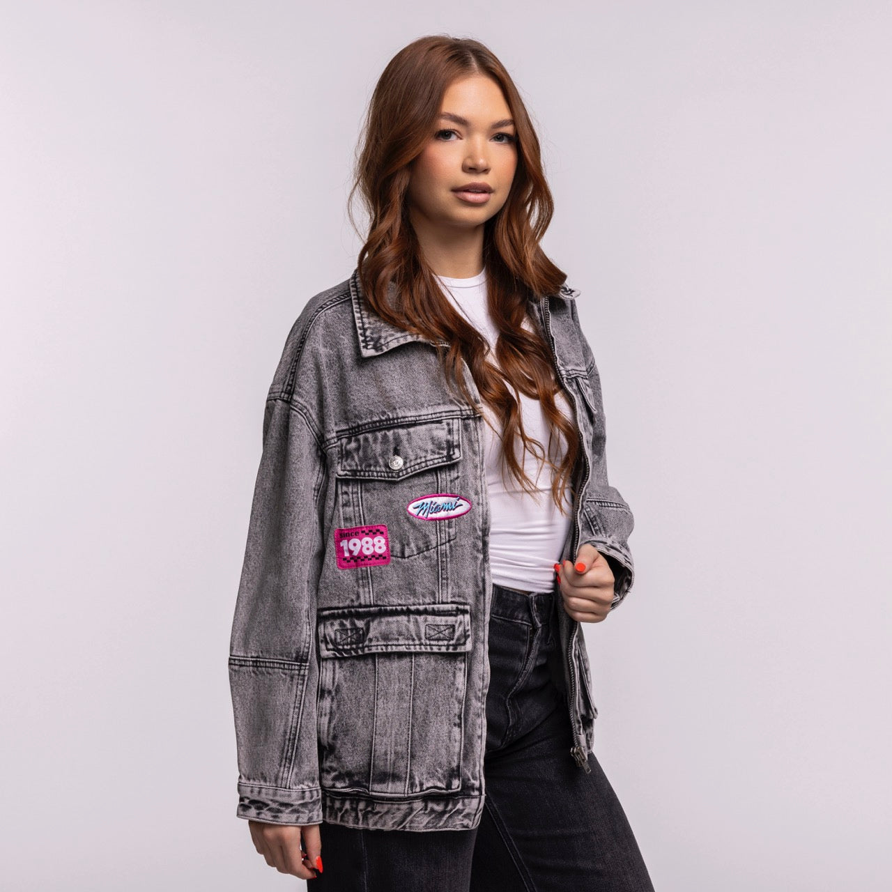 A young woman with long brown hair wears the Court Culture X The Wild Collective Original Vice Nights Denim Patch Unisex Jacket over a white shirt and black pants, standing against a plain light background.