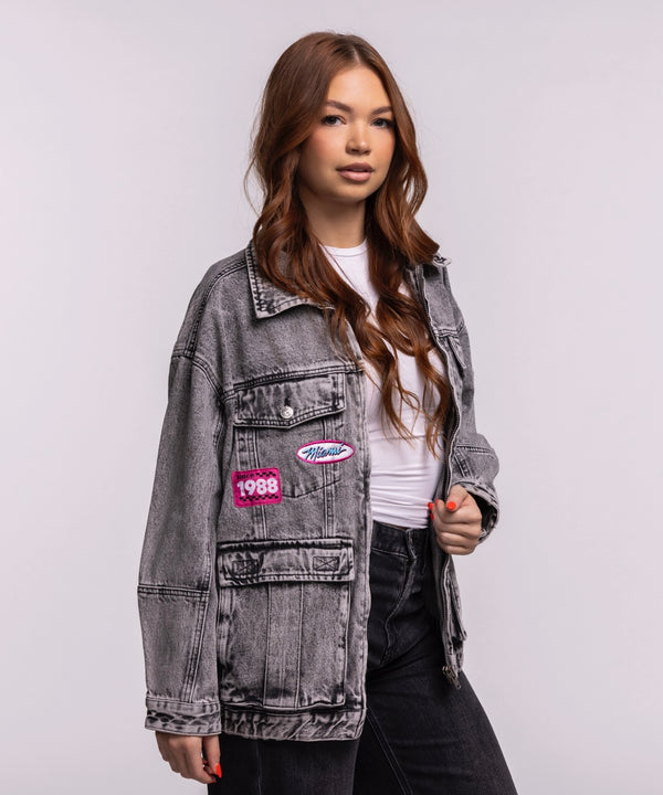 A young woman with long brown hair wears the Court Culture X The Wild Collective Original Vice Nights Denim Patch Unisex Jacket over a white shirt and black pants, standing against a plain light background.