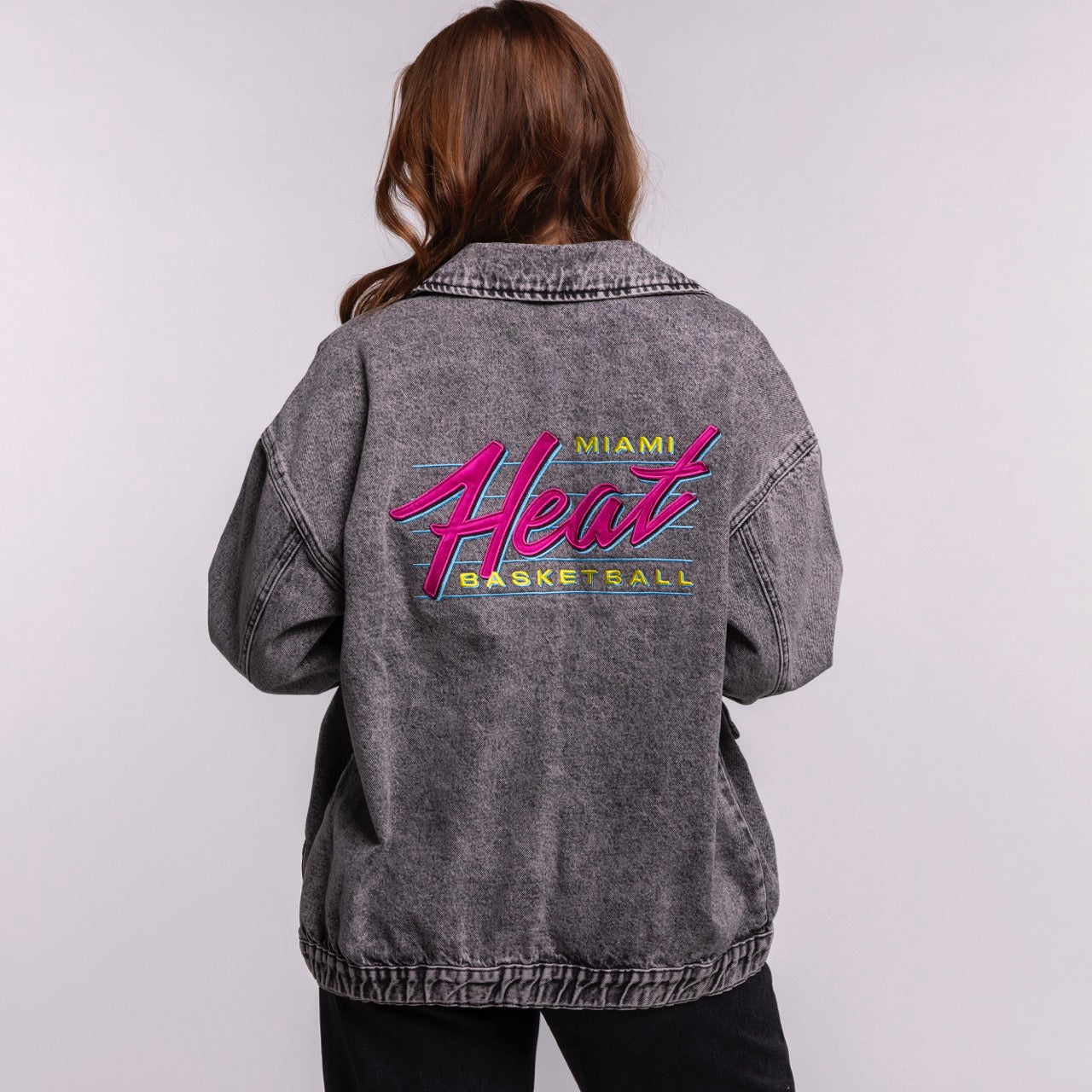 A person with long brown hair faces away, wearing the Court Culture X The Wild Collective Original Vice Nights Denim Patch Unisex Jacket, which features a colorful Miami Heat Basketball logo and stands out against a plain background.
