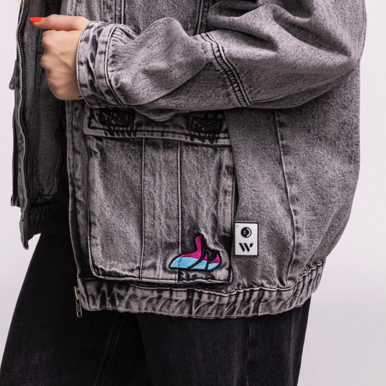 A person wears the Court Culture X The Wild Collective Original Vice Nights Denim Patch Unisex Jacket, featuring a colorful high-heeled shoe patch and logo tag. The gray jacket is standard fit, cotton, dry clean only; their left hand rests on it with black pants.