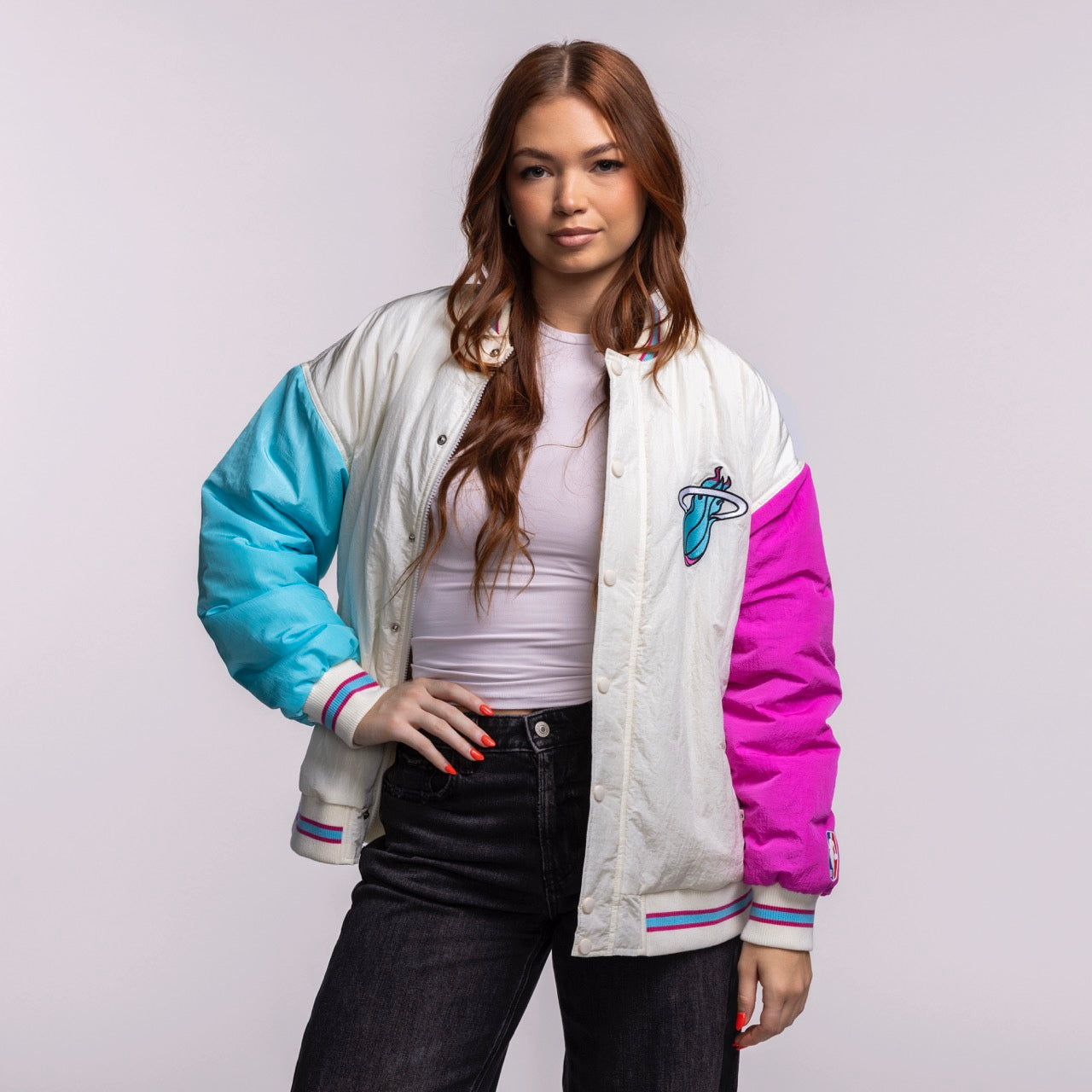 A woman with long brown hair models the Court Culture X Terez Original Vice Nights Unisex Bomber Jacket by Court Culture over a white top and black jeans, standing against a light gray background and looking at the camera with a relaxed expression.