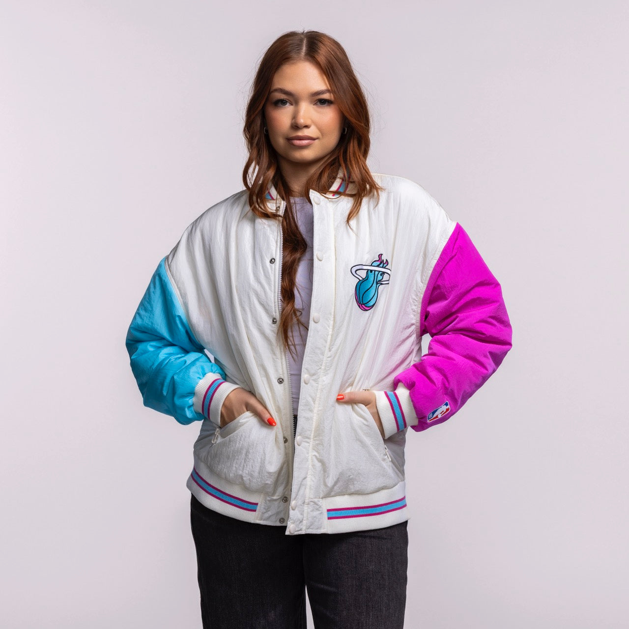 A woman with long brown hair wears the Court Culture X Terez Original Vice Nights Unisex Bomber Jacket by Court Culture, featuring blue and pink sleeves and a Miami Heat logo, over a light shirt and dark pants against a plain background.