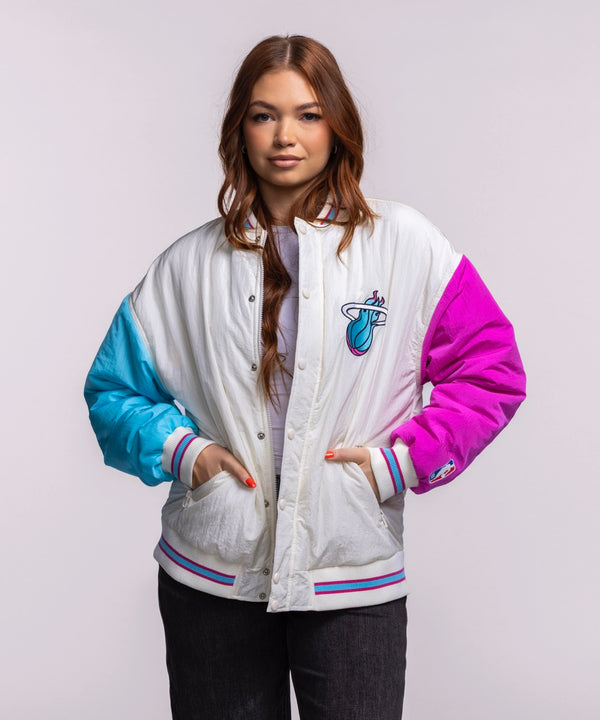 A woman with long brown hair wears the Court Culture X Terez Original Vice Nights Unisex Bomber Jacket by Court Culture, featuring blue and pink sleeves and a Miami Heat logo, over a light shirt and dark pants against a plain background.