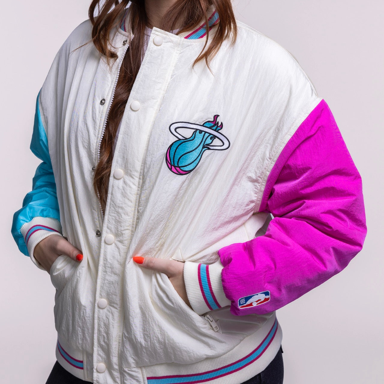 A person wears the Court Culture X Terez Original Vice Nights Unisex Bomber Jacket in white, pink, and teal. Only the lower half of their face is visible, hands are in pockets, and a flaming basketball logo appears on the chest.