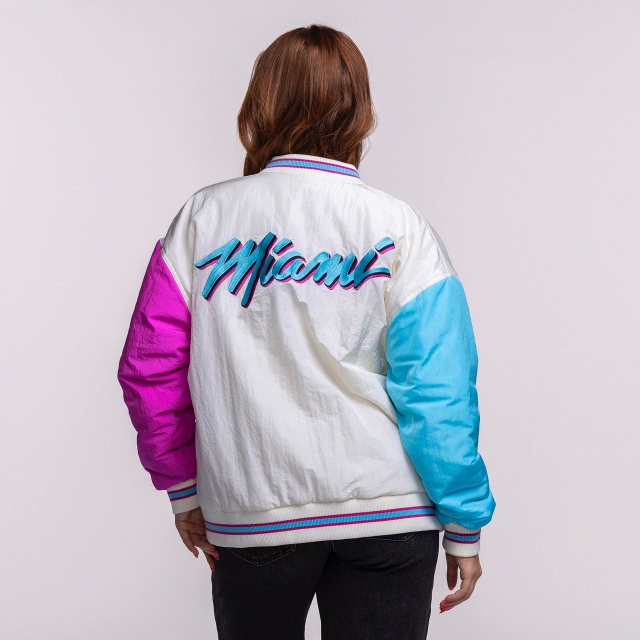 A person with long brown hair faces away, wearing the Court Culture X Terez Original Vice Nights Unisex Bomber Jacket by Court Culture, featuring “Miami” in retro script, a nylon shell, pink left sleeve, blue right sleeve, and striped collar and cuffs.