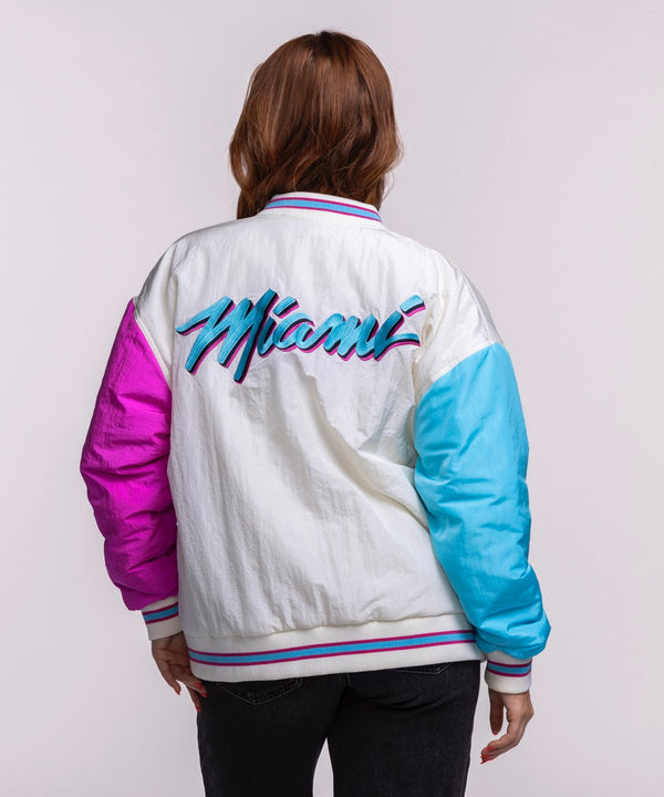 A woman with long brown hair wears the Court Culture X Terez Original Vice Nights Unisex Bomber Jacket by Court Culture, featuring blue and pink sleeves and a Miami Heat logo, over a light shirt and dark pants against a plain background.