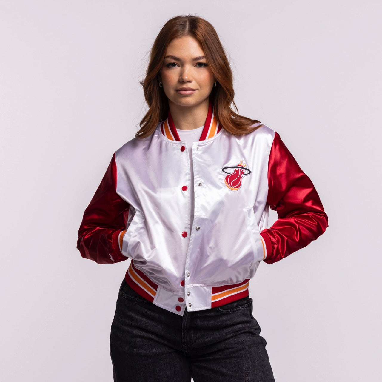 A woman stands confidently in the Mitchell & Ness Miami HEAT Women's Satin Jacket, featuring shiny red sleeves, a white cropped body, and a trendy varsity style—easy machine wash cold care pairs effortlessly with dark jeans.