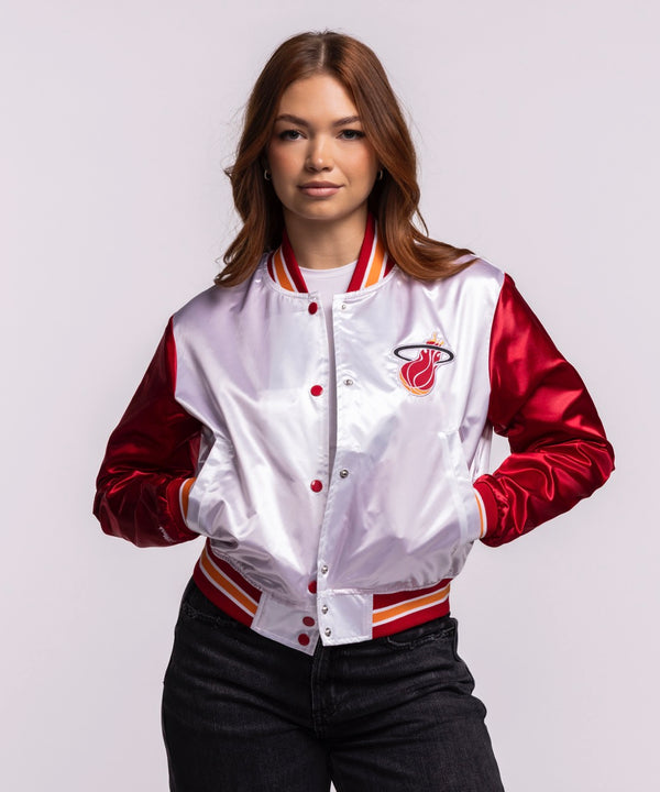 A woman stands confidently in the Mitchell & Ness Miami HEAT Women's Satin Jacket, featuring shiny red sleeves, a white cropped body, and a trendy varsity style—easy machine wash cold care pairs effortlessly with dark jeans.
