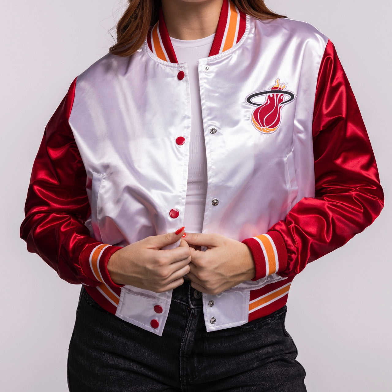 A woman wearing the Mitchell & Ness Miami HEAT Women's Satin Jacket in shiny red and white, styled cropped over a white shirt and black jeans, holds the jacket open to reveal the Miami Heat logo on the left chest.