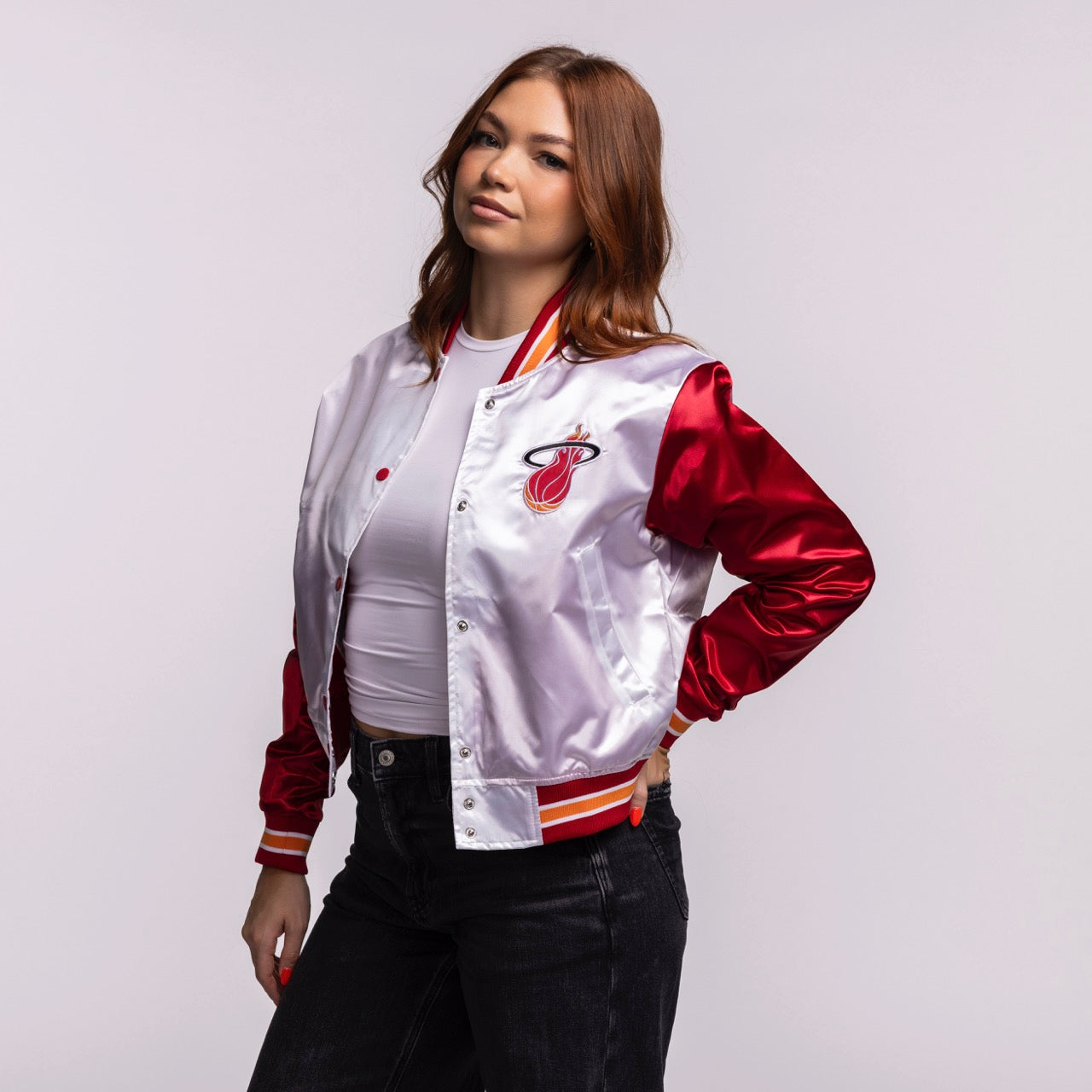 A woman with long brown hair models the Mitchell & Ness Miami HEAT Women's Satin Jacket in a stylish cropped fit over a white top and black jeans, posing with one hand on her hip against a plain light background.