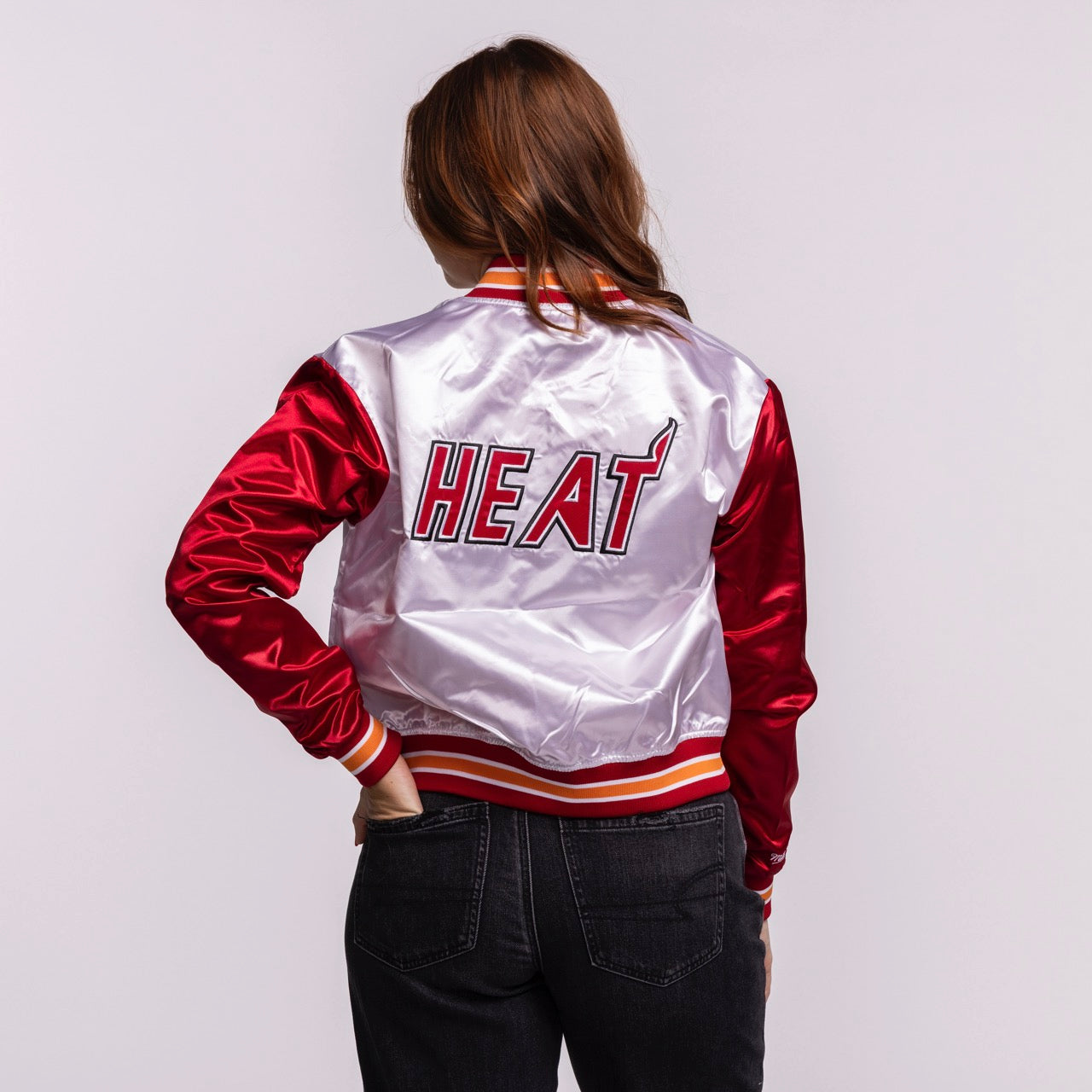 A woman with long brown hair, seen from behind, wears the Mitchell & Ness Miami HEAT Women's Satin Jacket—white and red polyester, 