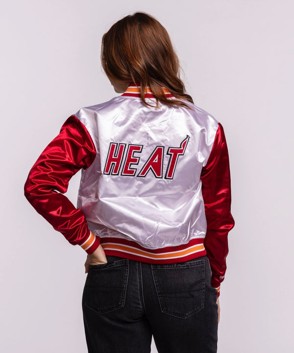 A woman stands confidently in the Mitchell & Ness Miami HEAT Women's Satin Jacket, featuring shiny red sleeves, a white cropped body, and a trendy varsity style—easy machine wash cold care pairs effortlessly with dark jeans.