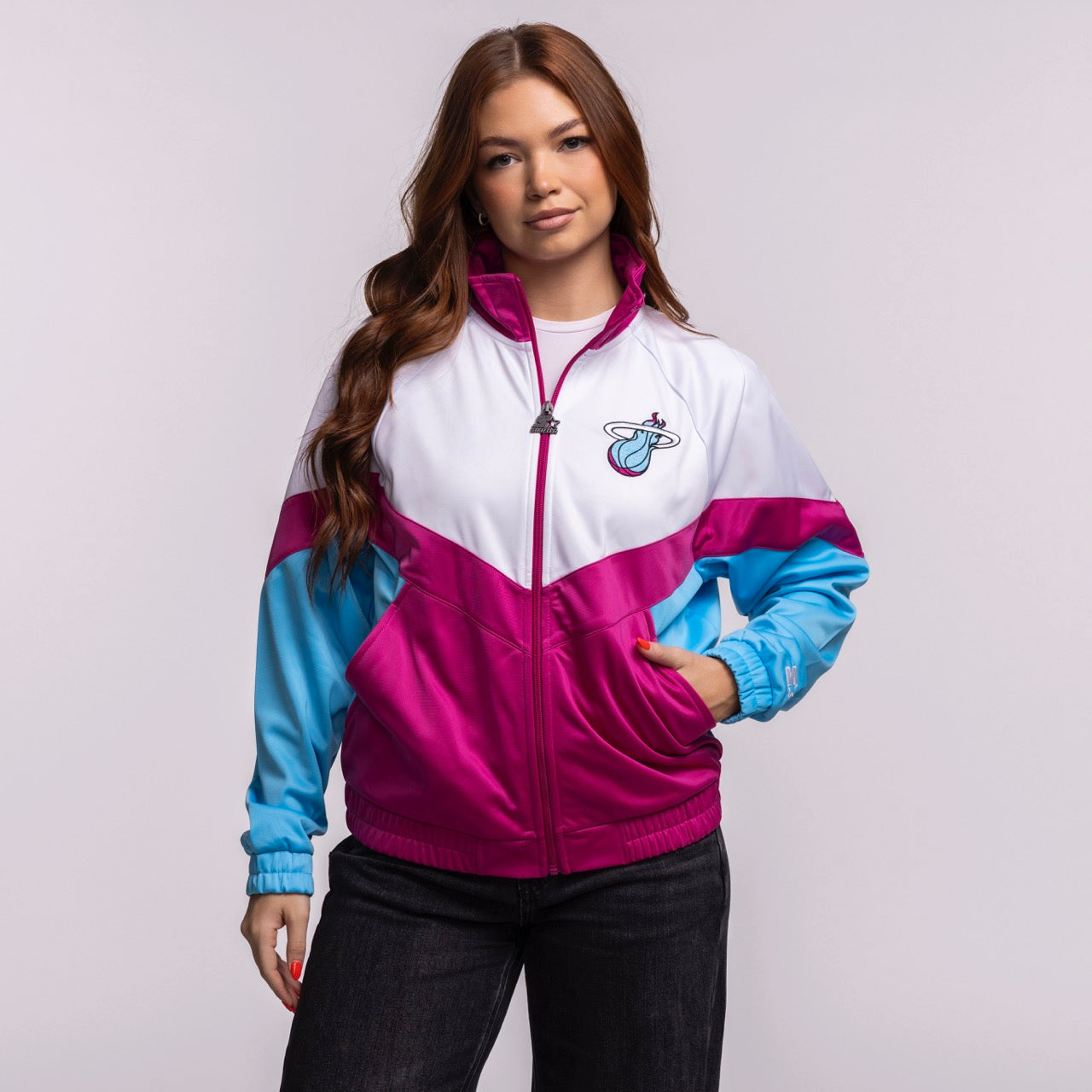 A woman with long brown hair wears the G-III Sports Miami HEAT Original Vice Nights Women's Jacket—white, pink, and blue with a team logo—paired with black pants against a plain light background. Machine wash cold for best care.