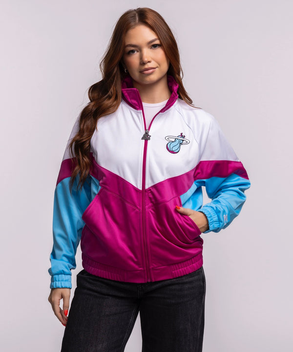 A woman with long brown hair wears the G-III Sports Miami HEAT Original Vice Nights Women's Jacket—white, pink, and blue with a team logo—paired with black pants against a plain light background. Machine wash cold for best care.