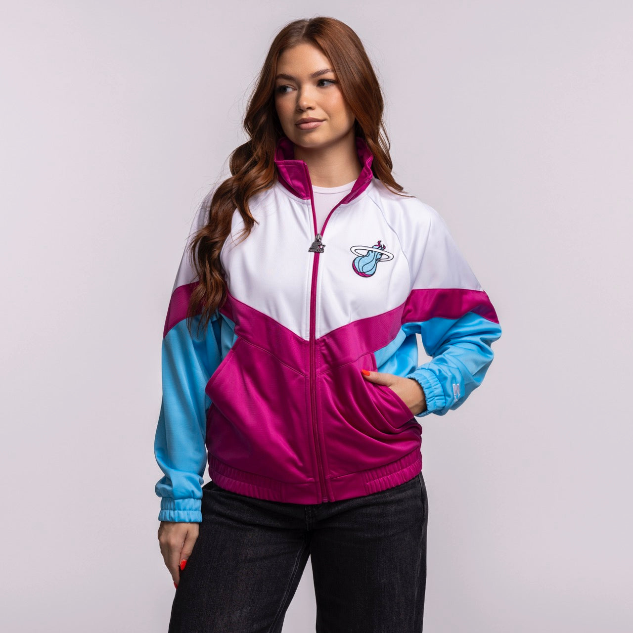 A woman with long brown hair wears the G-III Sports Miami HEAT Original Vice Nights Women's Jacket, a white, magenta, and blue polyester zip-up with a sports logo, standing casually with one hand in her pocket against a light gray background.
