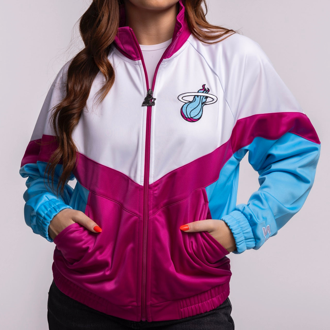 A woman sports the G-III Sports Miami HEAT Original Vice Nights Women's Jacket, featuring bold white, magenta, and blue colorblocks with the flaming basketball logo. This polyester jacket is machine washable—ideal for fans wanting a standout look.