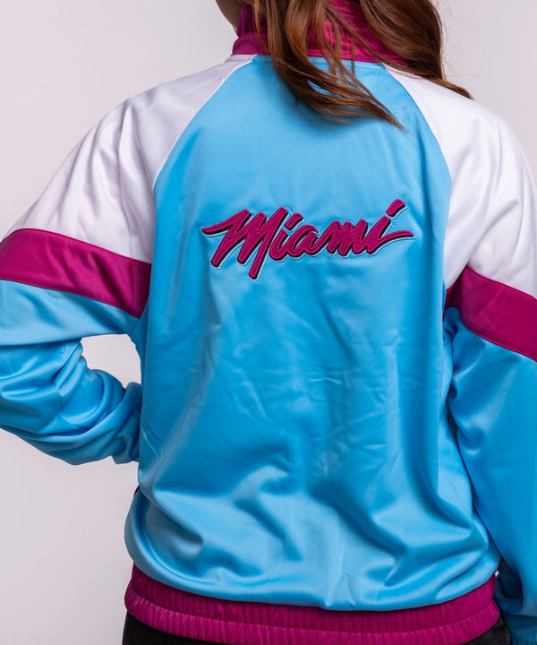 A woman with long brown hair wears the G-III Sports Miami HEAT Original Vice Nights Women's Jacket—white, pink, and blue with a team logo—paired with black pants against a plain light background. Machine wash cold for best care.