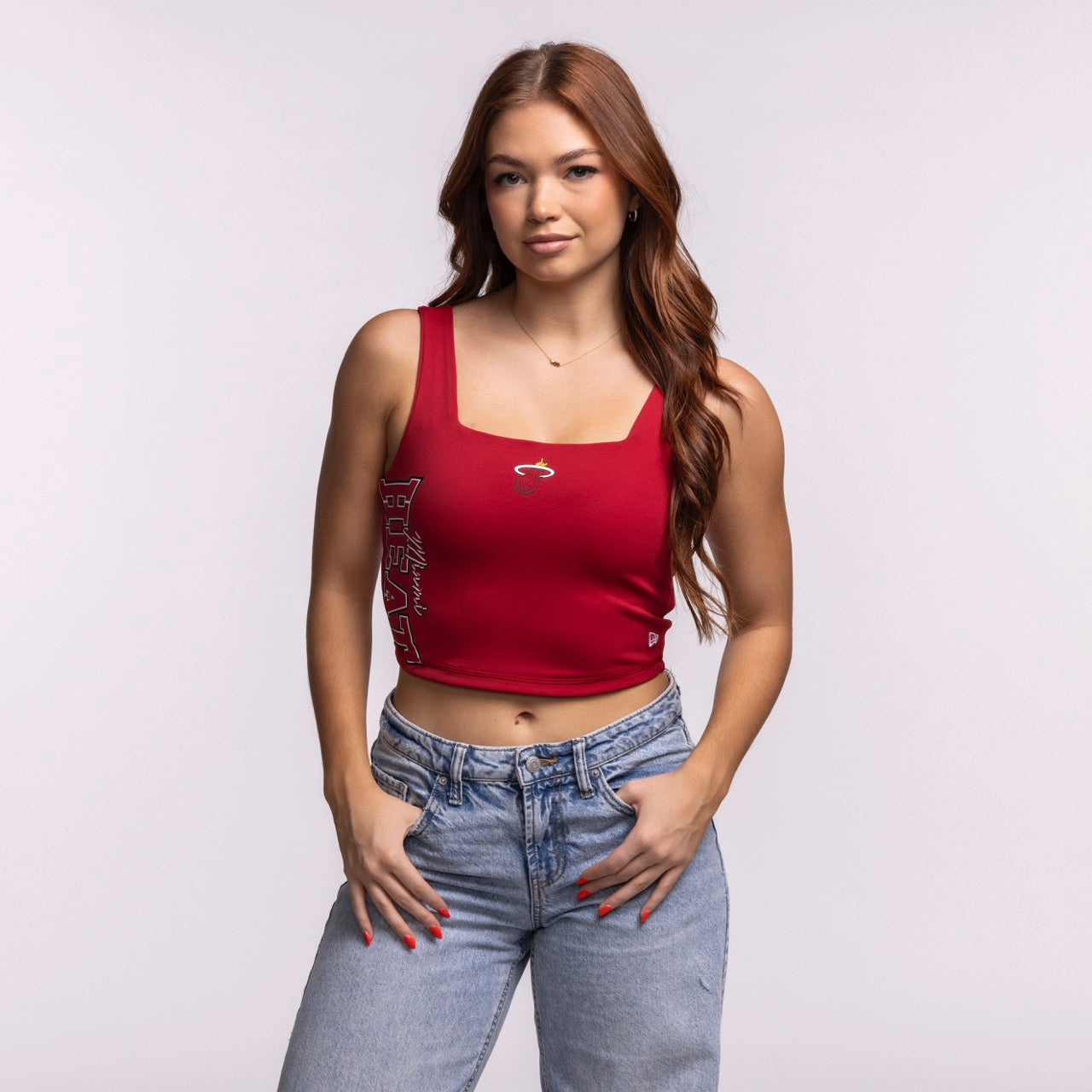A woman with long auburn hair wears the New Era Miami HEAT Reversible Women's Tank and light blue jeans, standing with hands on hips against a plain background—a stylish way to show team pride.
