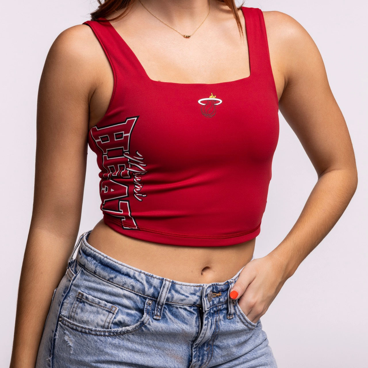 A woman wears a New Era Miami HEAT Reversible Women's Tank with a square neckline, featuring the team logo and name. She pairs it with high-waisted blue jeans and has orange-painted nails.