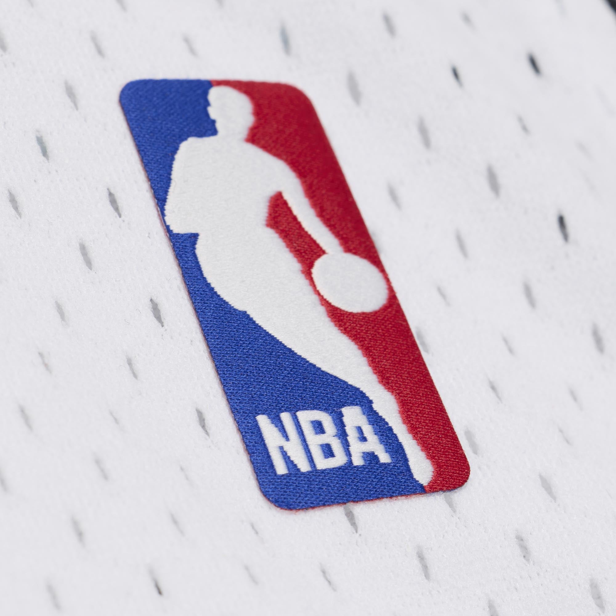 A red, white, and blue NBA logo patch stitched onto the white mesh polyester of a Dwyane Wade Mitchell & Ness Miami HEAT White Hot 2012-13 Swingman Jersey.