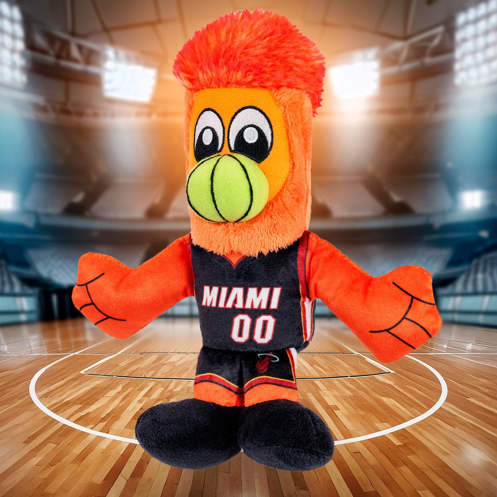 A Miami HEAT Burnie Icon Chibi Plushie by Bleacher Creatures, featuring orange fur, a green snout, and a MIAMI 00 jersey, stands on a basketball court inside a brightly lit indoor arena.