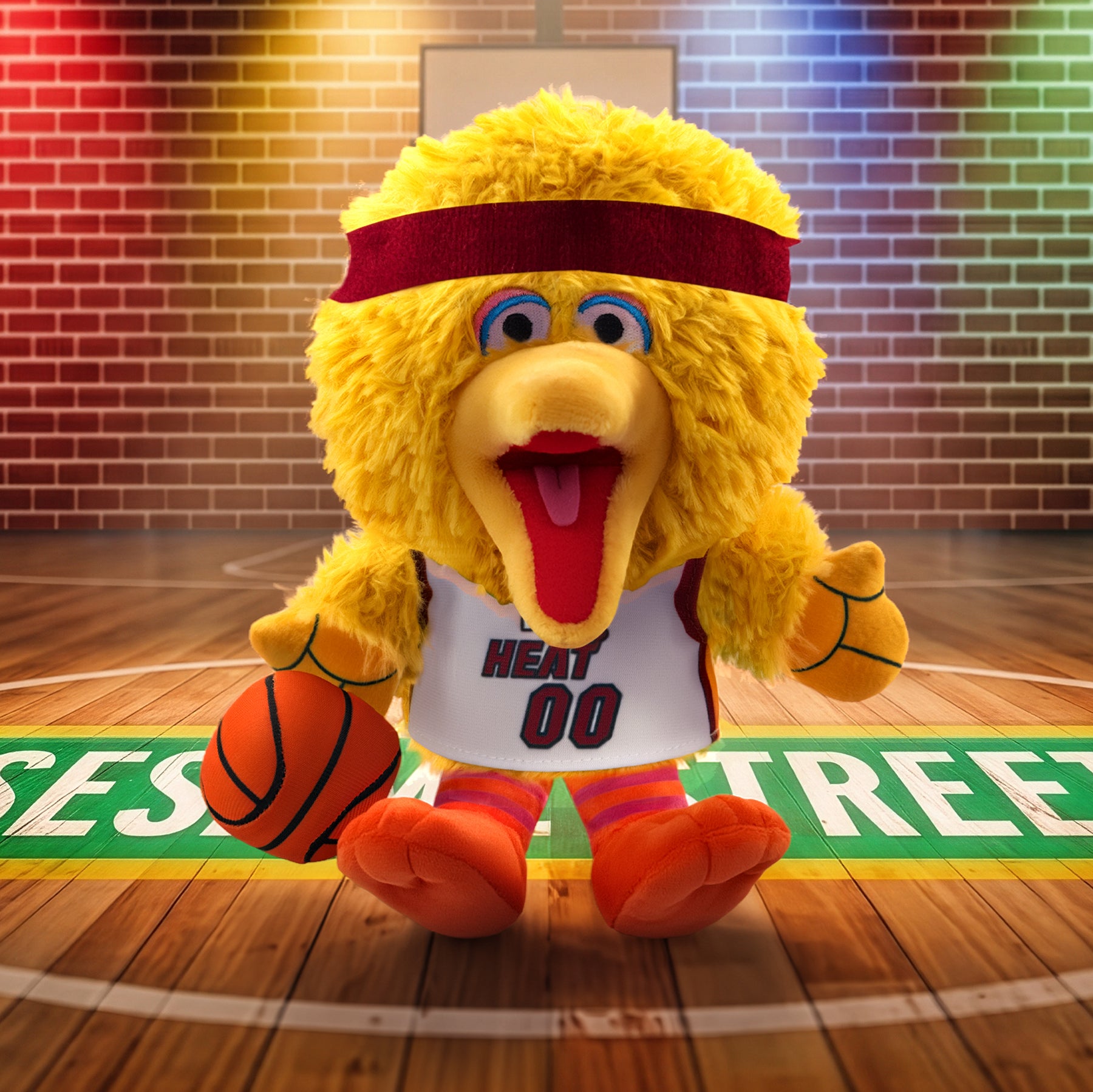 The Bleacher Creatures Miami HEAT Big Bird Association Plushie, featuring a plush yellow bird in a Miami HEAT 00 jersey holding a basketball, stands on a gym court with colorful lights highlighting the brick wall backdrop.
