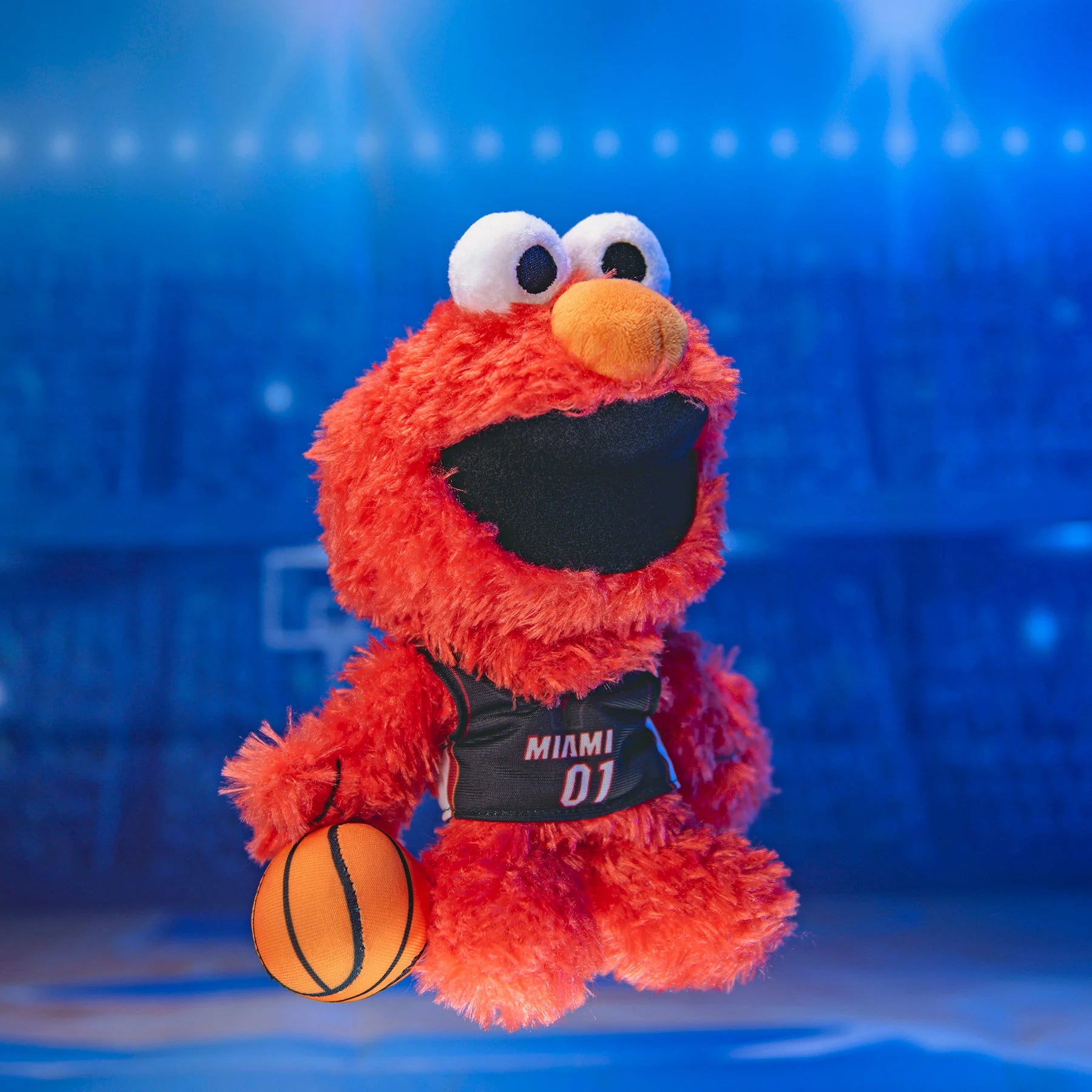 The Bleacher Creatures Miami HEAT Elmo Plush features Elmo in a black Miami 01 jersey, holding a basketball on a blue-lit court—a cute NBA x Sesame Street collectible for all ages.