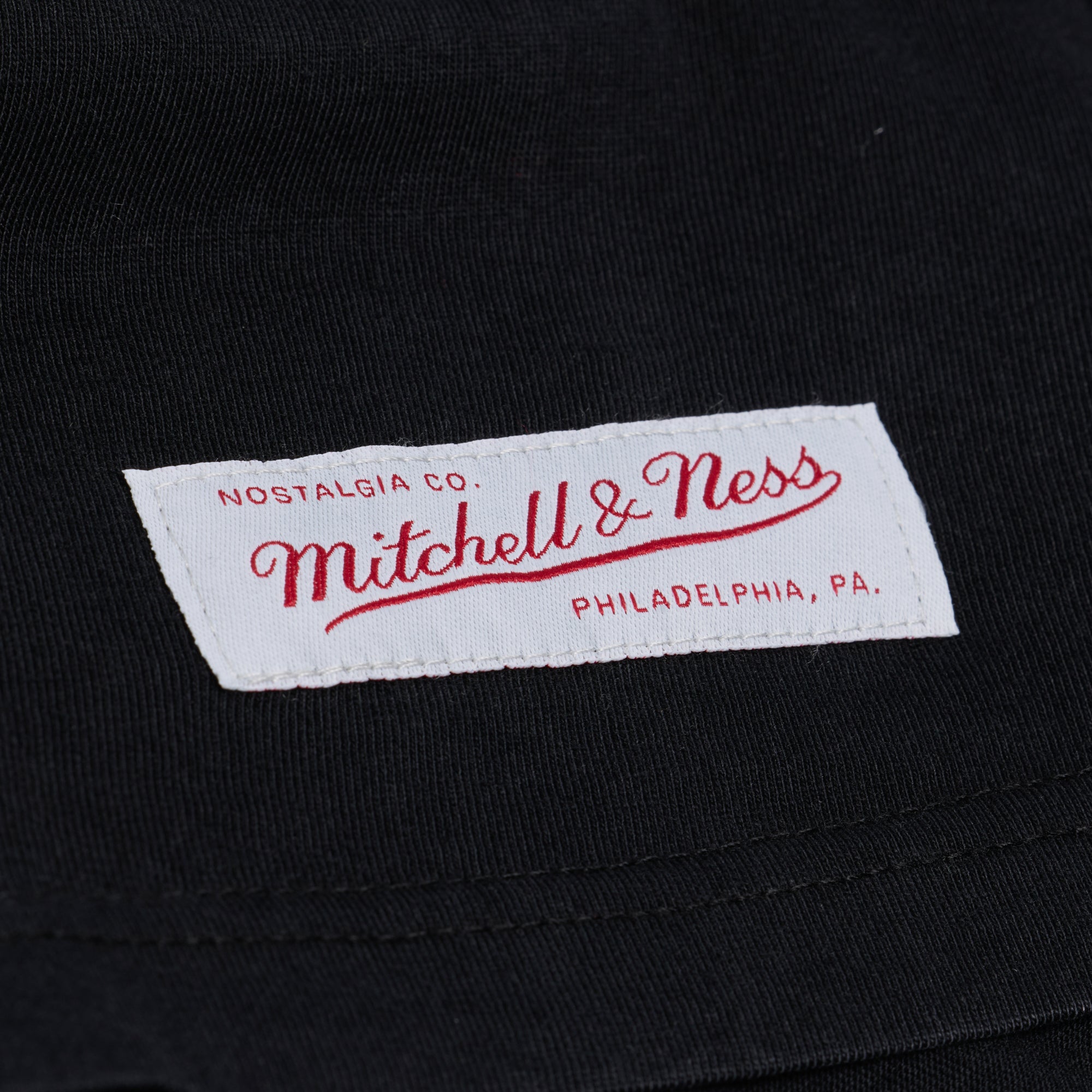 Close-up of a white Mitchell & Ness clothing label with red text, stitched onto a black fabric background. The label reads “Nostalgia Co. Mitchell & Ness Philadelphia, PA.” and is attached to a Mitchell & Ness Miami HEAT Core Essential Tee featuring subtle team graphic details.