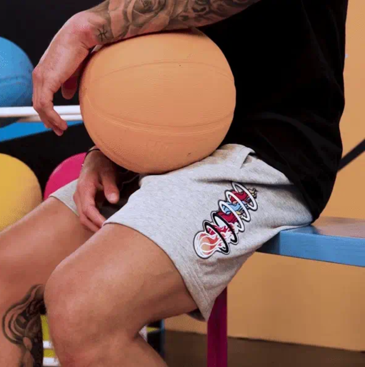 A tattooed individual sits on a bench, holding a basketball against their knee. They are wearing Court Culture Miami Mashup Vol. 2 88 Shorts—grey, 100% cotton shorts that feature a vibrant spiral design and an embroidered “88” patch. Other basketballs and a hoop can be seen in the background.