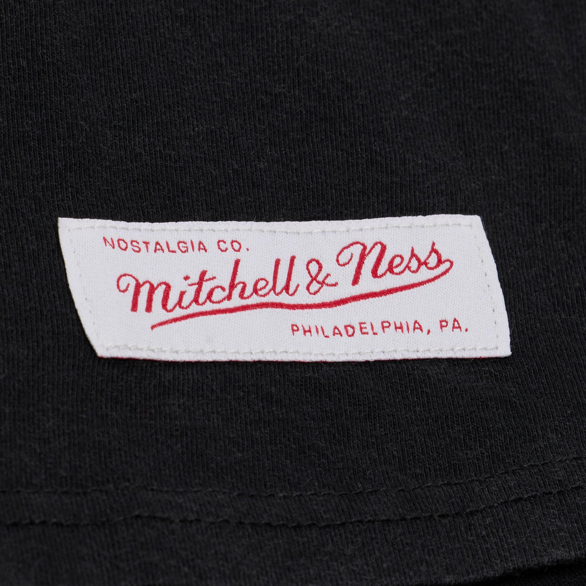 A white fabric label with red text that reads “Nostalgia Co. Mitchell & Ness Philadelphia, PA.” is sewn onto the black cotton Mitchell & Ness Miami HEAT Core Logo Women’s Crop Tee, a relaxed-fit garment designed for easy machine washing.