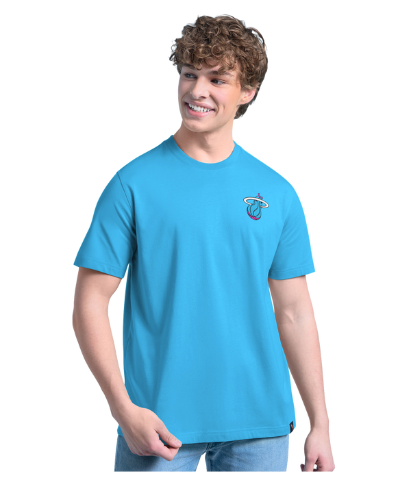A smiling young man with curly hair wears the G-III Sports Margaritaville Miami HEAT 'Everyday is Paradise' Tee and light blue jeans, standing against a plain white background.