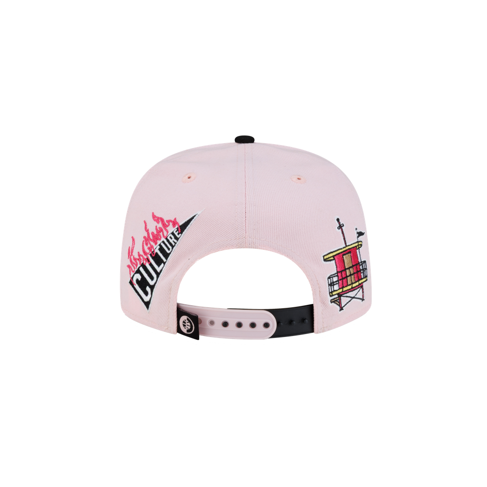 The Court Culture Original Vice Patch Snapback by Court Culture is a light pink polyester snapback cap viewed from the back, featuring a flaming Culture graphic on the left and a weather vane design with a red structure on the right. It sports a squared visor, slim crown, and includes a black adjustable strap for fit.