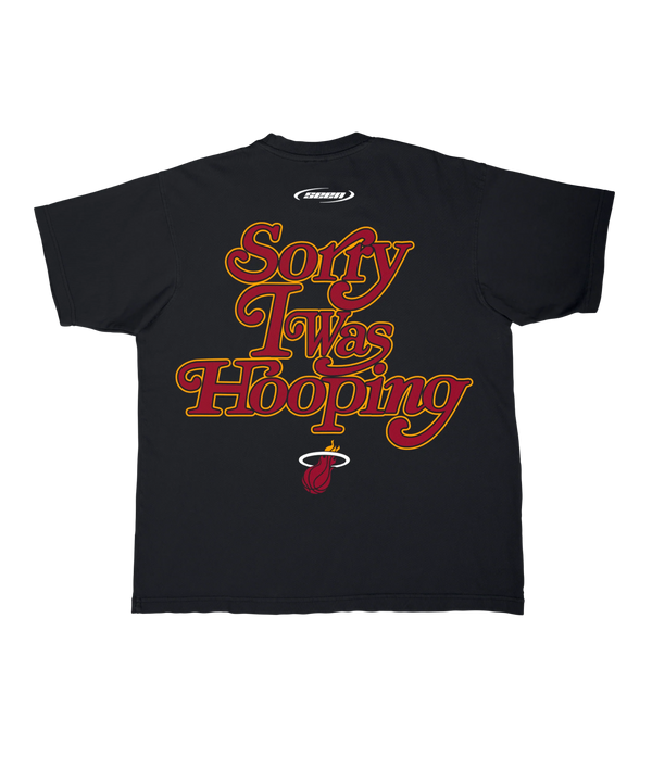 The Stadium Essentials Miami HEAT 'Sorry I Was Hooping' Tee is a black cotton shirt with the team logo on the left chest, shown on a white background. Machine wash cold to keep it comfortable and stylish.